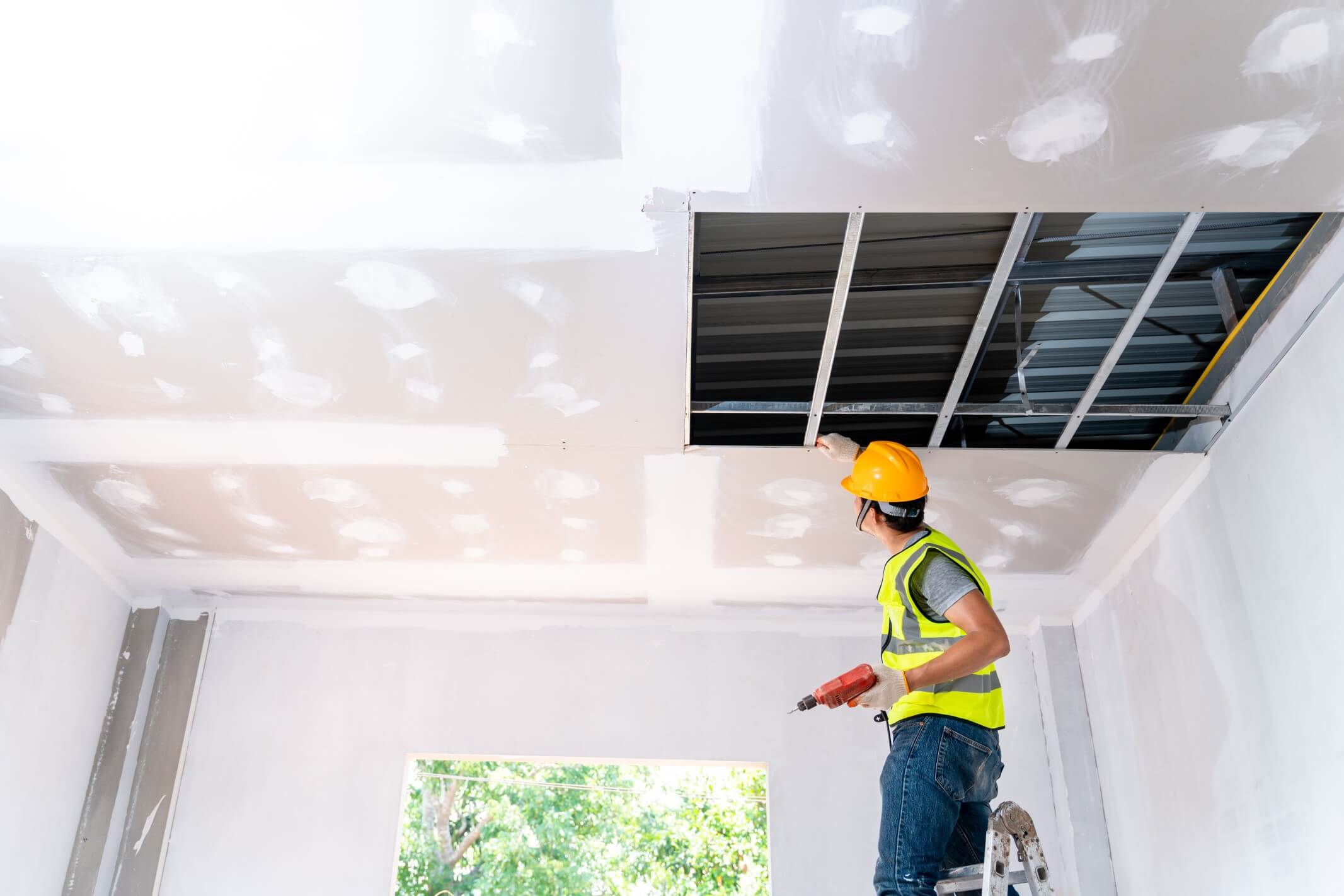 Construction worker assemble a suspended ceiling with drywall and fixing the drywall to the ceiling metal frame with screwdriver. Renovation, construction and ceiling installation concept.